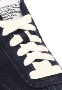 Zapatilla de navy oscuro con parte superior de textil, cordones blancos en contraste y acentos cosidos. Presenta una etiqueta visible en la parte superior con instrucciones de cuidado.