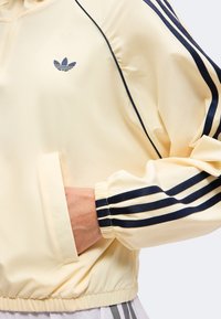 Απαλό κίτρινο μπουφάν Adidas με μαύρες τρεις ρίγες στα μανίκια, ελαστικά μανσέτες και τελείωμα και χέρι στην μπροστινή τσέπη.