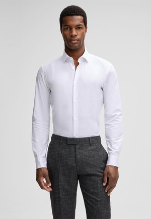 Mann in einem enganliegenden weißen Hemd mit Knöpfen und dunkelgrauen strukturierten Anzugshosen, steht vor einem schlichten hellen Hintergrund.