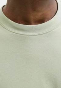 Tricou din bumbac de culoare verde deschis, cu guler rotund, având o textură netedă și cusături discrete, prezentat din perspectiva gâtului.
