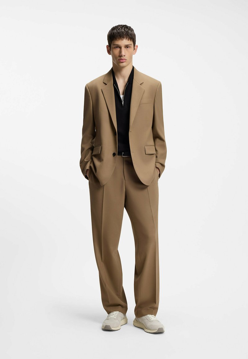 Jeune homme debout, les mains dans les poches, portant un costume beige, une chemise noire zippée et des baskets blanches sur fond blanc.