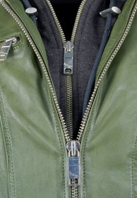 Grön läderjacka med silverfärgade dragkedjor, svart innerfoder och strukturerad yta. Detaljer inkluderar dubbla dragkedjor och sömnadsdetaljer.