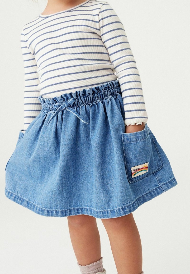 Enfant portant une jupe en denim bleu avec de grandes poches, un écusson portant l’inscription « Always Kind » avec un arc-en-ciel, et une chemise blanche à manches longues avec des rayures bleu marine.