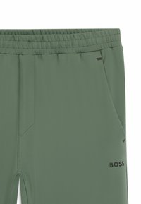 Green athletic pants with a smooth texture, elastic waistband, side pockets, and a small black logo on the leg. Features a tapered design.