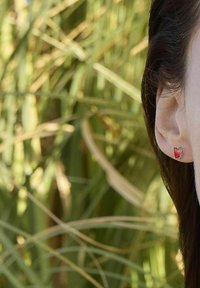 Primer plano de la oreja derecha de una persona que lleva un pendiente de gema roja rectangular, con cabello oscuro largo y un fondo de follaje verde difuminado.