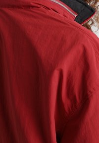 Červená bunda s viditelným zipem a černým límečkem, kterou nosí osoba s kudrnatými vlasy, zobrazená zezadu v úrovni ramen.