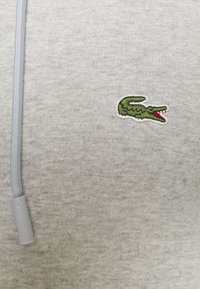 Felpa grigia con finitura tessurizzata, caratterizzata da un logo di coccodrillo ricamato in verde sul petto e una zip leggera di colore grigio chiaro.