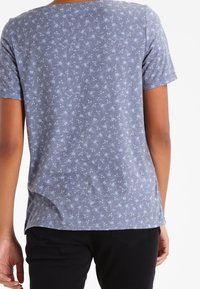 Top de manga corta en tejido azul grisáceo con un patrón floral. Cuello redondo y corte holgado, con una textura suave y dobladillo curvado.