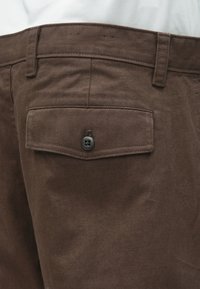 Brązowe spodnie materiałowe z tylną kieszenią zapinaną na guzik, teksturą jodełki oraz szlufkami na pasek wzdłuż talii.