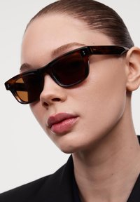 Ochelari de soare din material tartan, cu rame unghiulare, lentile întunecate și accente subtile din metal. Textură netedă cu un finisaj lucios.