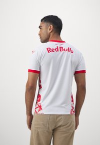 Bijela majica kratkih rukava s crvenim akcentima, tekst "Red Bulls" na leđima i uzorak na stranama. Glatka tekstura, atletski kroj.