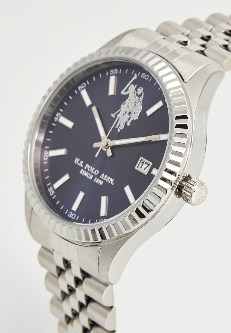Zilveren roestvrijstalen horloge met een donkerblauw dial, witte uurmarkeringen, datumweergave op 3 uur, en een gestructureerde bezel. Metalen schakelband.