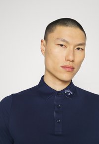 Navy polo shirt with a collared neck, button placket, and contrasting logo on the collar. Smooth fabric with a fitted silhouette.