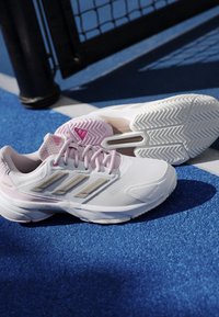 Hvide og lyserøde Adidas Courtjam sneakers placeret på en blå tennisbane nær netstolpen og den hvide grænselinje.