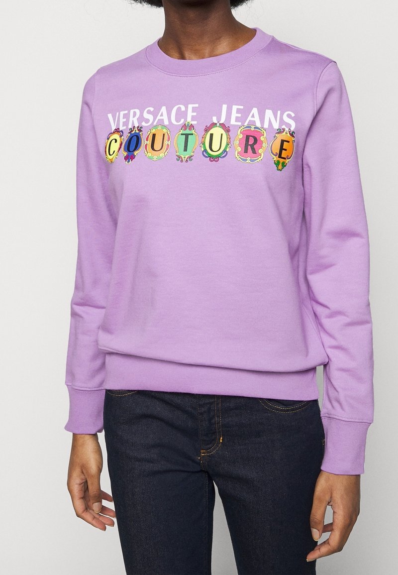 Sweat-shirt violet avec le texte "Versace Jeans Couture" et des motifs circulaires colorés. Présente des poignets côtelés et un col rond.