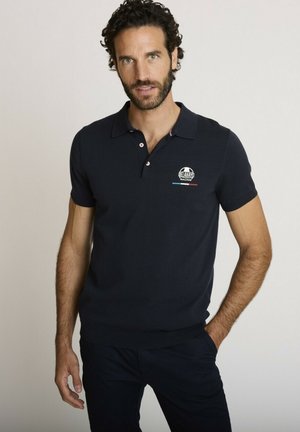 Homme aux cheveux bouclés et à la barbe portant un polo bleu marine avec le logo "Delahaye Racing", debout avec une main dans la poche.