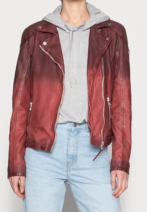 Osoba ubrana w czerwoną skórzaną kurtkę na szarej bluzie z kapturem i jasnoniebieskie, wysokiego stanu dżinsy, stojąca z rękami swobodnie opuszczonymi wzdłuż ciała.