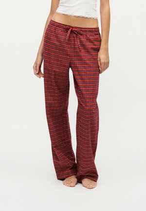 Femme portant un pantalon de pyjama à carreaux rouges et noirs avec une taille à cordon et un débardeur court blanc, debout pieds nus.