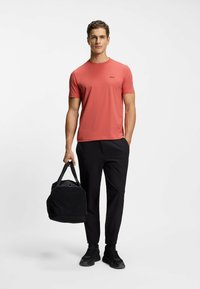 Coral short-sleeve T-shirt with black logo, paired with black trousers and black duffel bag. Model stands with a neutral background.