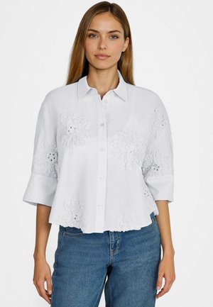 Frau trägt ein weißes Hemd mit Knöpfen und floralem Stickereimuster sowie blaue Jeans, steht vor einem schlichten weißen Hintergrund.