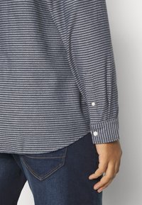 Camicia button-up a maniche lunghe con un motivo a righe blu e bianche, orlo curvo e polsini con un bottone, abbinata a jeans scuri.