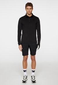 Musta pitkähihainen paita, jossa on neljännesvetoketju, yhdistettynä mustiin shortseihin. Valkoraidalliset sukat ja mustat urheilukengät viimeistelevät asun.