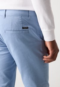 Helleblaue Baumwollhose mit glatter Textur, ausgestattet mit einer Gesäßtasche und einem kleinen Logotag, der auf der rechten Seite angenäht ist.