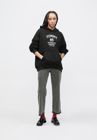 Čierna mikina s kapucňou s bielym grafickým nápisom, sivé džínsy a lesklé čierne topánky. Viditeľné vpletené vlasy a červené ponožky. Jednoduchý a neformálny outfit.