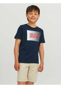 Tumši zils kokvilnas t-krekls ar baltu taisnstūra grafiku, kurā rakstīts "JACK & JONES" sarkanā krāsā. Apvienots ar sarullētiem bēšiem šortiem.