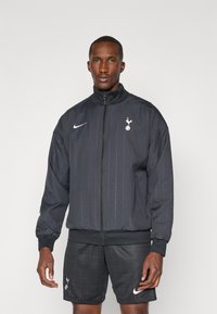 Sort træningsjakke med høj krave, lynlås og Nike swoosh. Har subtile spænder og Tottenham-logo på venstre bryst.