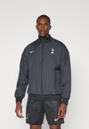 Giacca a track nera con colletto alto, chiusura con zip e logo Nike. Presenta sottili righe verticali e il logo del Tottenham sul lato sinistro del petto.
