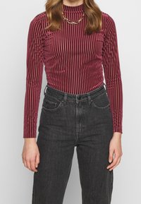 Burgundy ribbade långärmade topp med hög hals, med en texturerad elastisk väv och subtil glans, ihop med högmidjade svarta jeans.