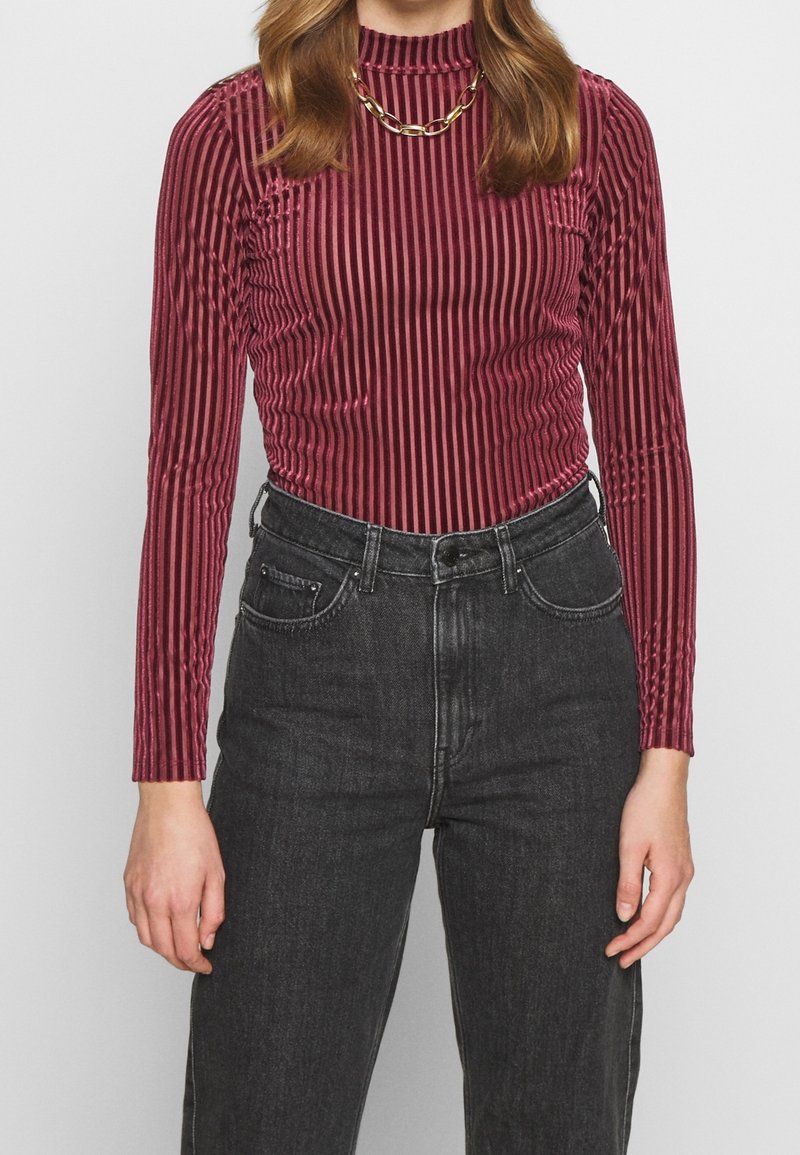 Burgundy ribbade långärmade topp med hög hals, med en texturerad elastisk väv och subtil glans, ihop med högmidjade svarta jeans.