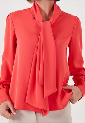 Kobieta ubrana w koralową bluzkę z długim rękawem z ozdobnym krawatem przy szyi oraz beżowe spodnie, z rękami spoczywającymi w okolicy talii.