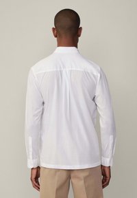 Camisa blanca de manga larga con cuello abotonado, textura suave y dobladillo recto. Presenta un yugo y puños con un solo botón.