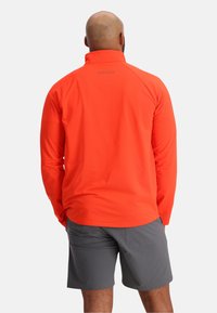 Oranje half-zip jack met een gestructureerd oppervlak, lange mouwen en een hoge kraag, gecombineerd met grijze shorts. Logo zichtbaar op de achterkant.