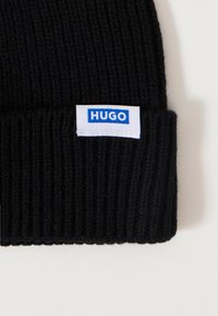 Crna pletena kapa s rebrastom teksturom. Ima bijelu oznaku s natpisom "HUGO" u plavim slovima. Klasičan, prilagođen dizajn za toplinu.