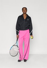 Μαύρο μπουφάν με φερμουάρ και λευκές λεπτομέρειες, συνδυασμένο με ροζ παντελόνι φαρδιάς γραμμής. Το μοντέλο κρατάει ρακέτα τένις και μπάλα τένις. Μαύρα παπούτσια.