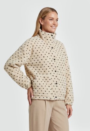 Kvinde smilende iført en beige fleecetrøje med små sorte prikmønstre, stående med den ene hånd på hoften, beige bukser.