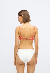 Czerwony biustonosz od bikini z wzorem, szerokimi ramiączkami i srebrnymi elementami, zestawiony z gładkimi, białymi, wysokim stanem spodniami kąpielowymi, widziany od tyłu.