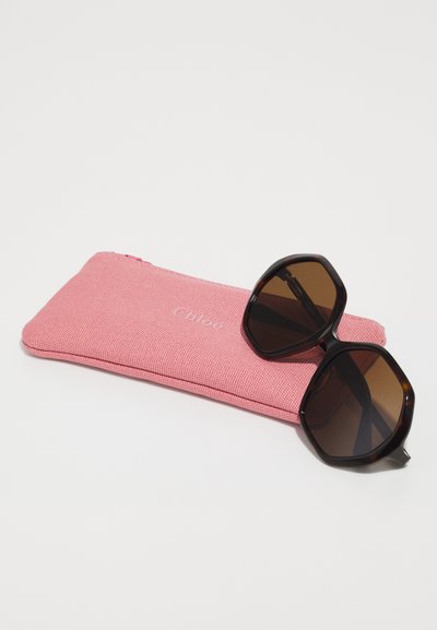 Črne osemkotne sončna očala z rjavimi lečami so postavljena na roza tkaninski etui s kamnom z vgraviranim imenom blagovne znamke "Chloé." Etui ima zadrgo.
