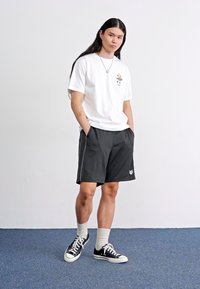 Hvit t-skjorte med kort erme og logo, svarte treningshorts med sidestriper, hvite ankelsokker og svarte joggesko. En enkel, uformell antrekk.