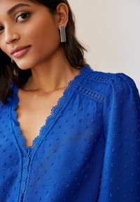 Modra bluza z teksturirano tkanino, ki ima pikčaste vzorce, valovit izrez in čipkaste poudarke spredaj ter na ramah.