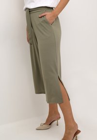 Olijfgroene, mid-length broeken gemaakt van zachte stof, met een ceintuur in de taille en zijsplitsingen, gecombineerd met beige hakken.