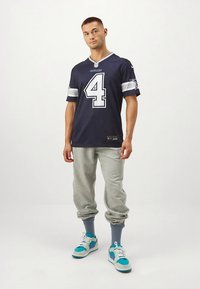 Marinblå Dallas Cowboys-tröja med vita ränder och nummer 4, kombinerad med grå sweatpants och flerfärgade sneakers.