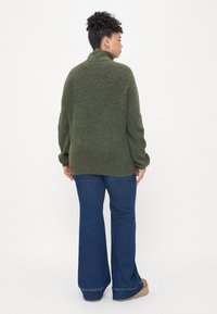 Suéter de punto verde con un cuello alto, con un acabado texturizado. Combinado con jeans oscuros de campana y calzado marrón claro.