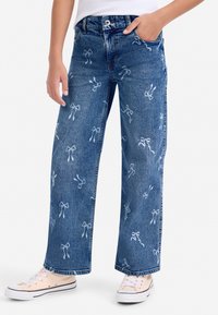 Denim jeans met wijde pijpen, voorzien van witte strikborduurwerk, voorzakken en een knoopsluiting. Gecombineerd met lichtgekleurde sneakers.