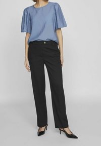 Blouse texturée bleu clair avec des manches bouffantes associée à un pantalon noir ajusté. Des chaussures à talons hauts noires complètent la tenue.