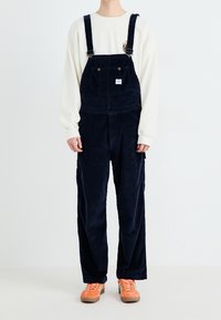 Marinefarbene Corduroy-Latzhose mit verstellbaren Trägern, Fronttaschen und einem kleinen Logo-Patch, kombiniert mit einem cremefarbenen Shirt und orangenen Schuhen.