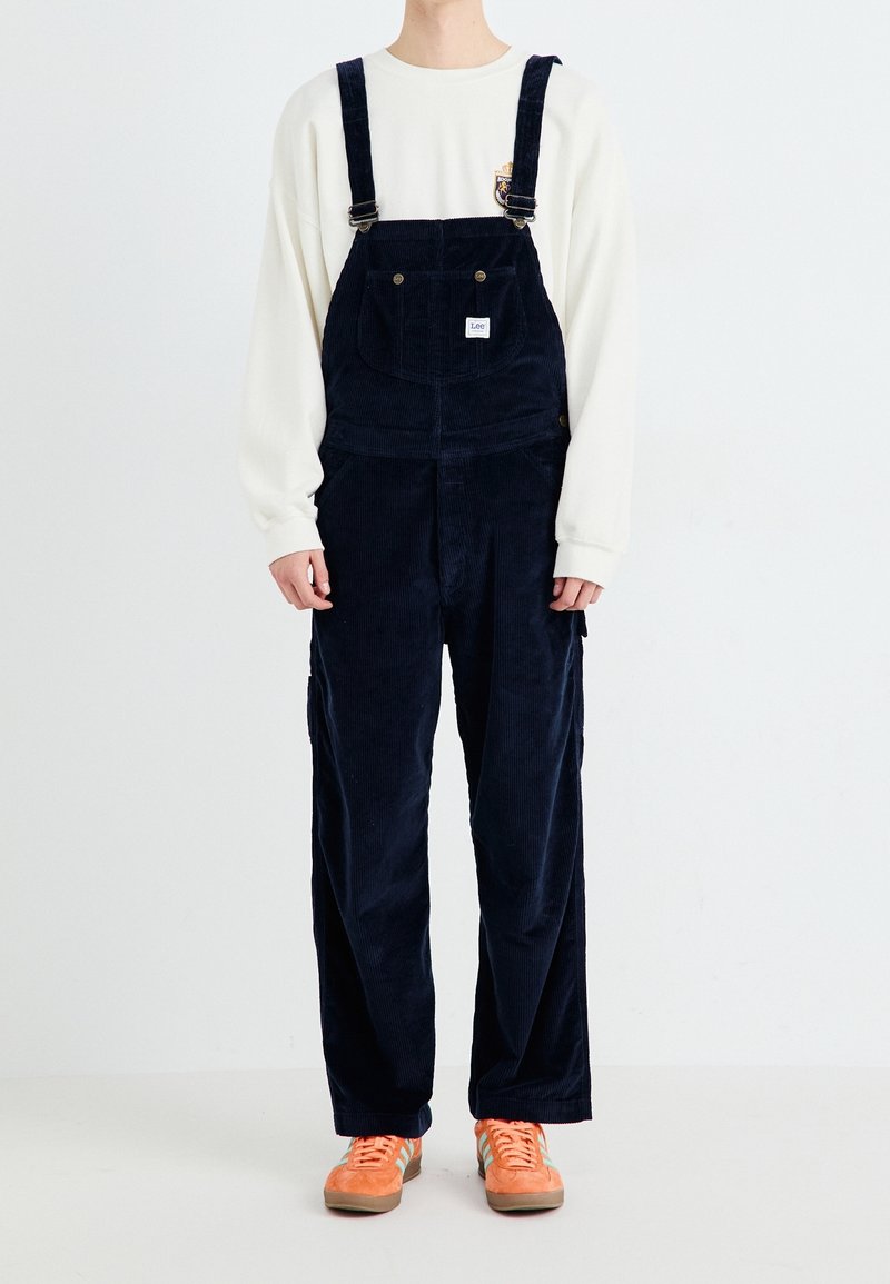 Marinefarbene Corduroy-Latzhose mit verstellbaren Trägern, Fronttaschen und einem kleinen Logo-Patch, kombiniert mit einem cremefarbenen Shirt und orangenen Schuhen.